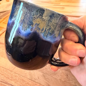 May include: A handmade ceramic mug with a glossy finish. The mug features a dark blue base that transitions to lighter blue and gold tones near the rim. The handle is a matching dark blue. The mug is held over a wooden surface.