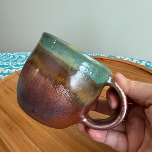 May include: A handmade ceramic mug with a unique color gradient. The mug transitions from a deep reddish-brown at the base to a teal green at the rim. The handle is a matching brown. The mug is on a wooden surface.