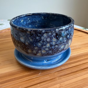 May include: A round ceramic planter with a deep blue glaze featuring a mottled, crystalline texture in shades of light blue and white. The bowl-shaped vessel sits securely on a matching circular blue saucer with a smooth, glossy finish.