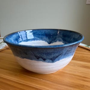 May include: A large ceramic bowl featuring a two-tone design with a glossy deep blue glaze on the upper portion and rim, transitioning into a matte white finish on the lower half. The bowl has a wide, circular opening and a smooth, rounded base.