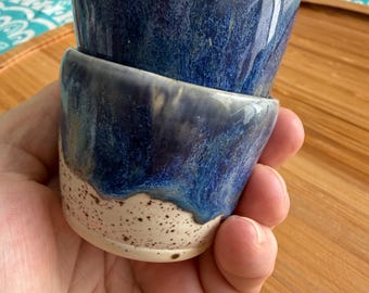 May include: Two handmade ceramic cups stacked. The cups have a dark blue glaze with lighter blue and white speckles. The bottom portion of the cups is a speckled off-white color.