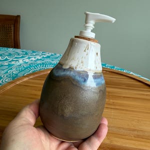 May include: A handmade ceramic soap dispenser with a white pump. The dispenser has a brown base, a blue-grey midsection, and a white top. The dispenser is held in a hand and sits on a wooden tray.