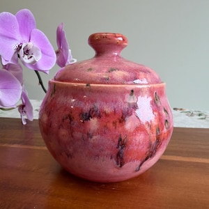 May include: A round ceramic lidded jar featuring a glossy glaze in shades of pink, coral, and magenta with dark, mottled accents. The jar has a smooth, curved body and a matching lid with a small, rounded knob handle on top for easy removal.