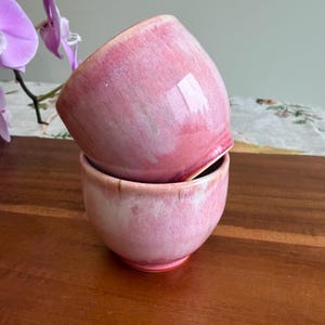 May include: Two ceramic cups stacked vertically, featuring a glossy glaze with blended shades of pink, white, and soft coral. The cups have a rounded, handleless shape with a smooth finish, showcasing a gradient of color from deep pink to pale white.