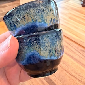 May include: Two small, square ceramic bowls stacked together. The bowls have a dark blue base that fades into a lighter blue and tan color. The bowls are held in a person's hand.