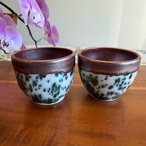 May include: Two ceramic cups featuring a rounded shape with a smooth, dark metallic brown glaze on the upper rim and a white base decorated with a mottled, dark green and black crystalline pattern that creates a distinct, textured visual effect.