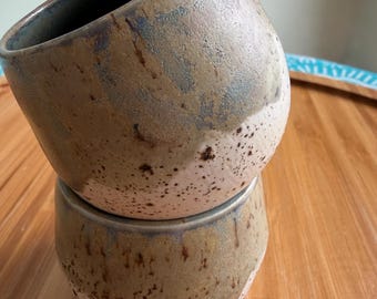 May include: Two handmade ceramic mugs stacked on top of each other. The mugs have a speckled, textured surface with a color gradient of brown, beige, and blue. The interior of the top mug is dark brown.