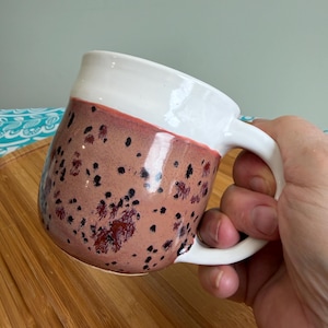 May include: A handmade ceramic mug with a white top and a speckled, reddish-brown body. The mug features a curved handle and is held by a person's hand. The speckled design includes dark spots, adding visual interest to the mug's surface. The mug is on a wooden surface.
