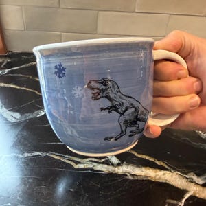 May include: A blue ceramic mug with a white handle and rim. The mug features a black illustration of a Tyrannosaurus Rex and a snowflake. The mug is held by a person.