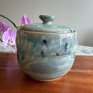 May include: A round ceramic jar with a matching lid, featuring a glossy glaze in shades of blue, teal, and grey. The surface displays a mottled, textured finish with darker blue speckles and a smooth, rounded knob handle on the lid.