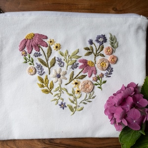 May include: White canvas zippered pouch with an embroidered floral heart design. The heart is filled with pink, yellow, and white flowers, along with green leaves. A cluster of pink hydrangea flowers is in the foreground.