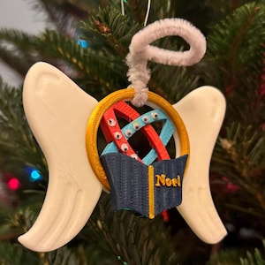 Könnte beinhalten: Weisser Engel Flügel Anhänger mit einem goldenen Kreis, der ein rotes, blaues und weißes Design enthält. Der Kreis hat ein blaues Buch mit dem Wort "Noël" darauf.