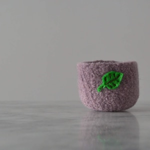 May include: A small, purple, fuzzy planter with a green felt leaf attached to the side.