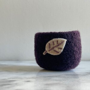 May include: A small, dark purple felt bowl with a beige felt leaf appliqué. The bowl is sitting on a white marble surface.