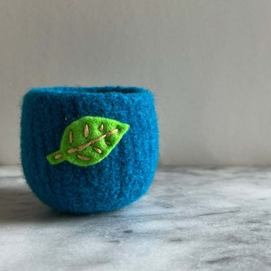 May include: A teal blue felt planter with a green leaf embroidered on the side.