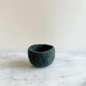 May include: A small, dark teal felt bowl. The bowl is round with a slightly textured surface and a dark interior. The bowl is sitting on a white and gray marble surface.