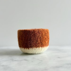 以下が含まれることがあります： 手作りの小さなフェルトボウル。上部は暖かく赤みがかった茶色で、下部はクリーム色です。ボウルは丸みを帯びた形状で、質感のある表面があり、居心地の良い自然な美しさを醸し出しています。明るい色の表面に置かれています。