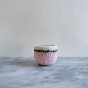 Puede incluir: Un pequeño recipiente redondo tejido a mano. El recipiente es principalmente rosa claro, con una parte superior blanca y una fina raya negra y gris. El artículo está sobre una superficie de mármol.