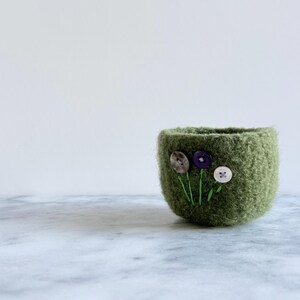 Può includere: Una piccola ciotola rotonda in feltro verde oliva con tre steli verdi ricamati sormontati da tre bottoni distinti: uno grigio, uno viola e uno bianco. La ciotola ha una superficie testurizzata e soffice con un design semplice e aperto.