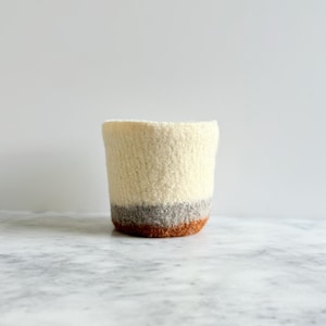 Puede incluir: Un portavasos cilíndrico hecho a mano de lana afieltrada. El portavasos es principalmente de color crema, con una banda gris y marrón cerca de la base. El portavasos está diseñado para sostener una taza o un recipiente pequeño. El portavasos mide aproximadamente 10 cm de alto.