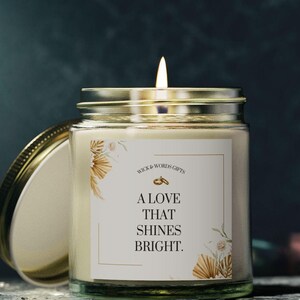 Puede incluir: Una vela blanca en un tarro de vidrio con tapa dorada. La etiqueta en el tarro dice "A love that shines bright." con un diseño de anillo dorado.