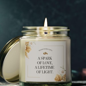 Könnte beinhalten: Ein klares Glas-Kerzengefäß mit einem weißen Etikett, auf dem "A spark of love, a lifetime of light." steht. Das Etikett hat ein goldenes Ringdesign und florale Illustrationen. Die Kerze ist angezündet und die Flamme ist sichtbar.