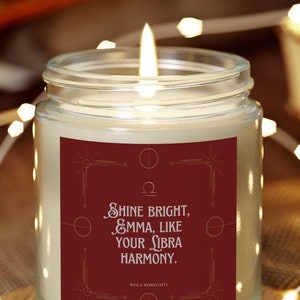 May include: A glass jar candle with a red label that reads "Shine bright, Emma, like your Libra harmony." The candle is lit and surrounded by white string lights.
