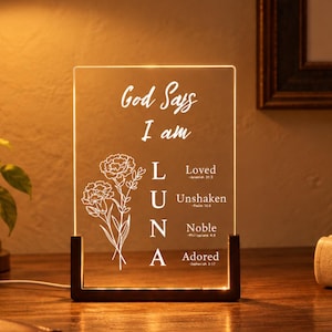 May include: A rectangular clear acrylic plaque with a dark wooden base, illuminated from below. The plaque features the text "God Says I am" above the name "LUNA" vertically, with "Loved," "Unshaken," "Noble," and "Adored" beside it, and a floral line drawing.