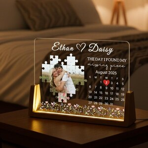 May include: A personalized illuminated acrylic photo frame with a wooden base. The frame features a photo within a puzzle design, the names "Ethan & Daisy", and the text "THE DAY I FOUND MY missing piece August 2025". The frame measures approximately 8 inches tall.
