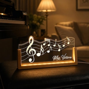 May include: A clear acrylic light display featuring a musical staff with a treble clef and various musical notes, illuminated by a warm light base made of light-colored wood. The text Miss. Victoria is etched onto the acrylic surface.