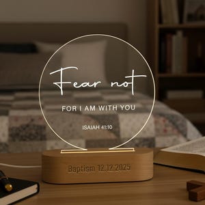 May include: A lighted, circular sign with the words "Fear not" and "For I am with you" in white script. The sign sits on a wooden base with the date "Baptism 12.12.2025" engraved. A Bible and cross are also visible.
