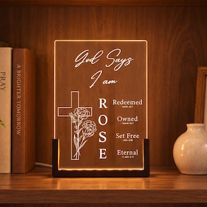 May include: A rectangular acrylic light plaque featuring the text God Says I am ROSE with a cross and flower illustration. The vertical text ROSE aligns with the words Redeemed, Owned, Set Free, and Eternal, each accompanied by a Bible verse reference.
