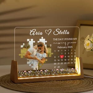 May include: A personalized acrylic photo display with a wooden base. The display features a photo of a couple kissing, puzzle pieces, and the names "Aria" and "Stella." It includes a calendar for February 2025 and the phrase "THE DAY I FOUND MY missing piece."