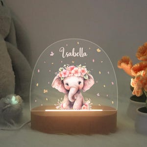 Könnte beinhalten: Ein klares Acryl-Nachtlicht mit einem Holzsockel. Das Nachtlicht zeigt einen rosa und weißen Elefanten mit einer Blumenkrone, umgeben von rosa und weißen Blumen. Der Name "Isabella" ist auf der Vorderseite aufgedruckt.