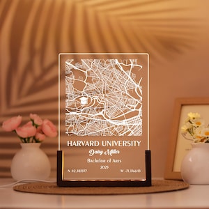May include: A rectangular clear acrylic plaque with a white engraved map of Harvard University, featuring a graduation cap icon. The text reads Harvard University, Daisy Miller, Bachelor of Arts, 2025, and coordinates N 42.583577 W -71.136645, set in a dark wooden base.