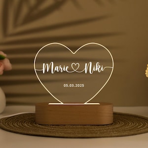May include: A heart-shaped LED lamp with the names "Marie & Niki" and the date "05.03.2025" etched on the illuminated surface. The lamp sits on a wooden base, creating a warm, ambient glow. The overall design is simple and elegant.