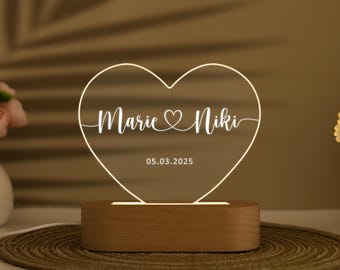 May include: A heart-shaped LED lamp with the names "Marie & Niki" and the date "05.03.2025" etched on the illuminated surface. The lamp sits on a wooden base, creating a warm, ambient glow. The overall design is simple and elegant.