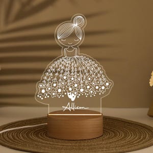 May include: A decorative night light featuring a line drawing of a girl in a dress, with the name "Allison" inscribed. The light is made of clear acrylic and sits on a wooden base. The girl's dress is adorned with floral details.
