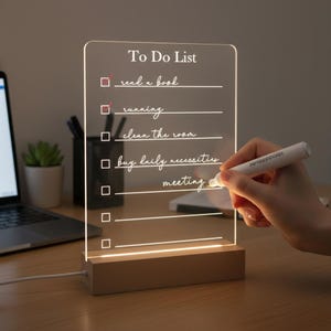 May include: A clear acrylic To Do List illuminated by a warm light, with a wooden base. The list includes tasks like "read a book" and "running." A hand is writing on the list with a white marker. The text "To Do List" is at the top.
