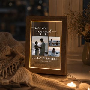 Op de afbeelding: Een rechthoekige houten lijst met geïntegreerde ledverlichting, met drie foto's van een koppel op een transparant paneel. De tekst "we're engaged" staat bovenaan, met "JULIAN & ISABELLE" en "04.08.2026" onderaan afgedrukt.