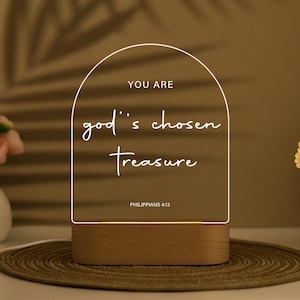 May include: A decorative LED night light with a wooden base and a clear acrylic panel. The panel features the text "YOU ARE god's chosen Treasure" and "PHILIPPIANS 4:13" in white script. The light emits a soft glow.