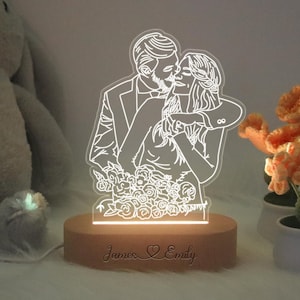 Op de afbeelding: Een houten basis met een gepersonaliseerde inscriptie "James & Emily" en een witte omtrek van een kussend stel op een transparant acryl paneel. Het stel is omringd door bloemen.