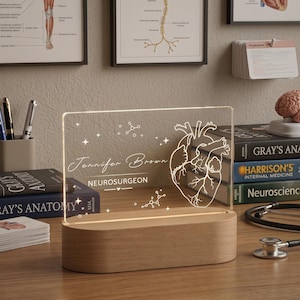 Puede incluir: Una luz nocturna LED personalizada con una base de madera. El panel acrílico transparente presenta una ilustración detallada de un corazón anatómico, el nombre "Jennifer Brown" y el título "NEUROSURGEON". Estructuras moleculares decorativas y estrellas también están grabadas en el panel.
