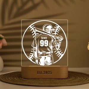 May include: A baseball-themed night light with a wooden base. The light features a white graphic of a baseball player with the name "Scott" and the number "99" on the jersey. The base reads "Est. 2025".