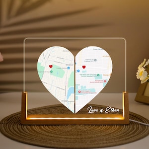 May include: A heart-shaped map night light with two halves, each displaying a different location marked with a red heart. The base is made of wood and has a warm, glowing light. The names "Juna & Ethan" are written in cursive.