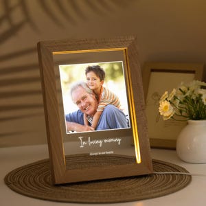 Op de afbeelding: Een houten fotolijst met een warme gloed verlicht een foto van een man en een jongen. De lijst heeft een witte inscriptie met de tekst "In liefdevolle herinnering, Altijd in onze harten".