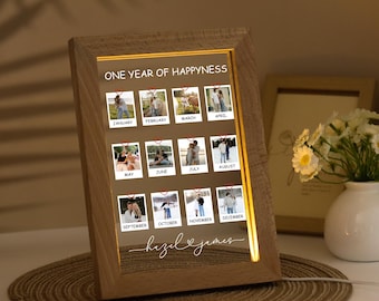 May include: A wooden framed photo display with the text "ONE YEAR OF HAPPYNESS" and twelve small photos representing each month. The names "hazel & james" are written in cursive at the bottom. The frame has a warm, glowing light.