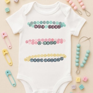 May include: White baby onesie with the text "I'VE BEEN A-LONG TIME COMING" in a colorful beaded design. The onesie is surrounded by baby accessories, including safety pins and a pacifier clip with beads. A cute and playful design.