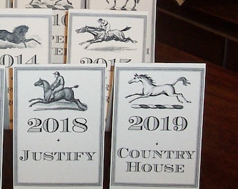 May include: A set of five white cards with black text and illustrations of horses. The cards are numbered 2014, 2015, 2018, 2019, and 2017. The cards also have the names "Justify" and "Country House" printed on them.