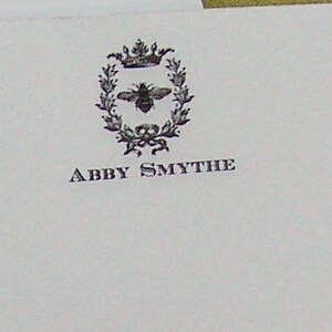 May include: A black and white image of a bee with a crown on its head inside a wreath. The text "Abby Smythe" is below the wreath.